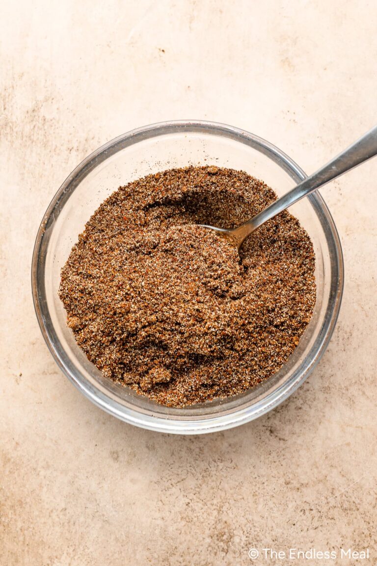 A glass bowl filled with a brown, granular coffee rub spice mix and a metal spoon rests on a light-colored surface.