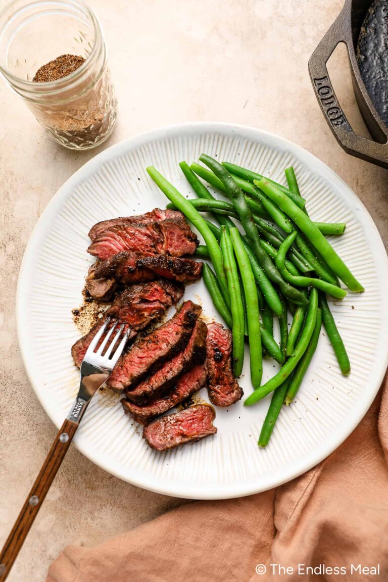 A white plate with Coffee Rubbed Steak slices and green beans, a fork resting on the steak. Nearby, there’s a glass jar of seasoning and a cast iron skillet, hinting at the flavorful preparation.