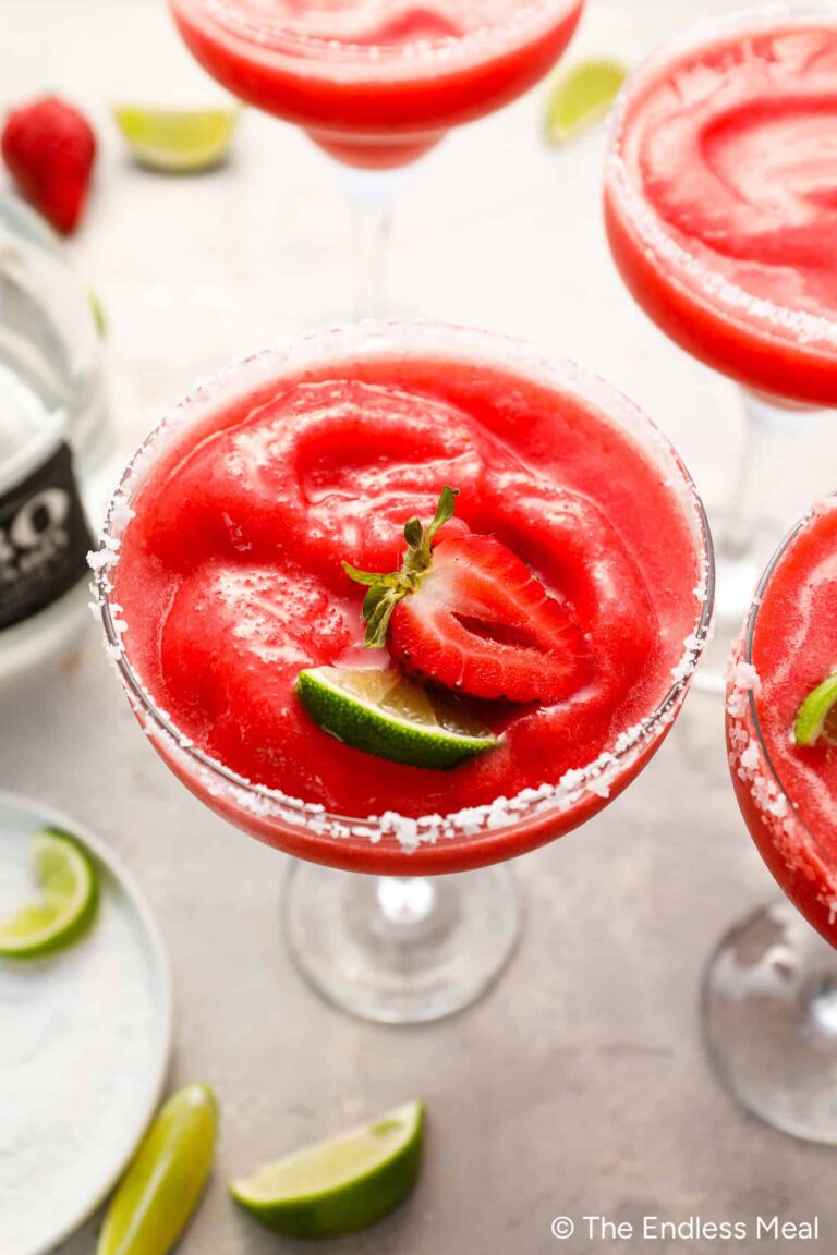 A salt-rimmed glass filled with a vibrant Frozen Strawberry Margarita, garnished with a strawberry slice and lime wedge. Other similar drinks and lime wedges are nearby, enhancing the lively atmosphere.