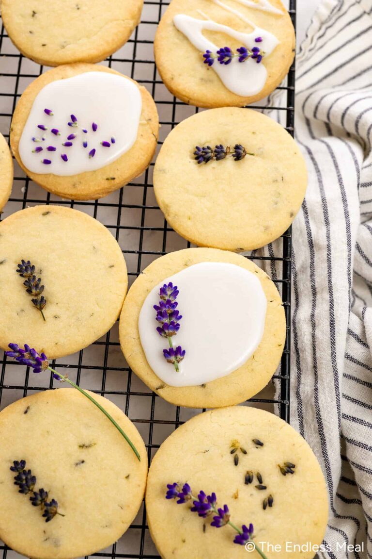 Round Lavender Shortbread Cookies, some topped with white icing and garnished with lavender flowers, are cooling on a black wire rack beside a striped cloth.