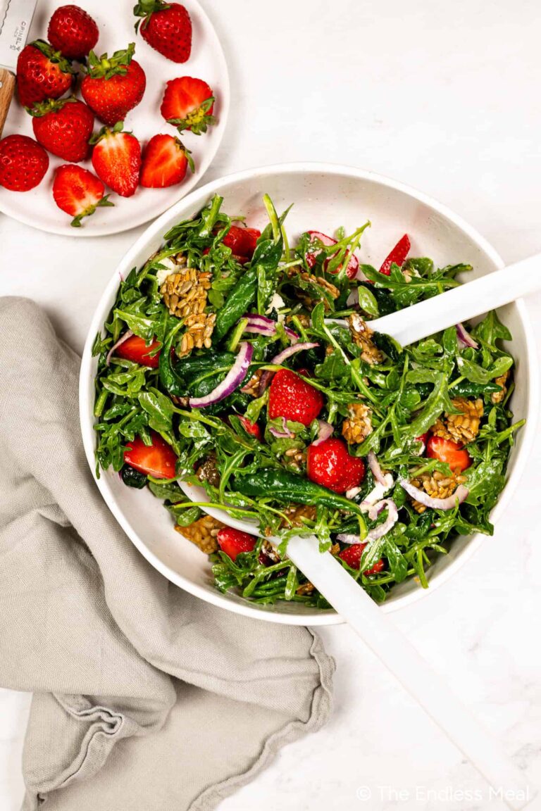 A bowl of Strawberry Arugula Salad with strawberries, red onions, sunflower seeds, and vinaigrette. A plate of halved strawberries and a gray napkin are beside the bowl on a white surface.