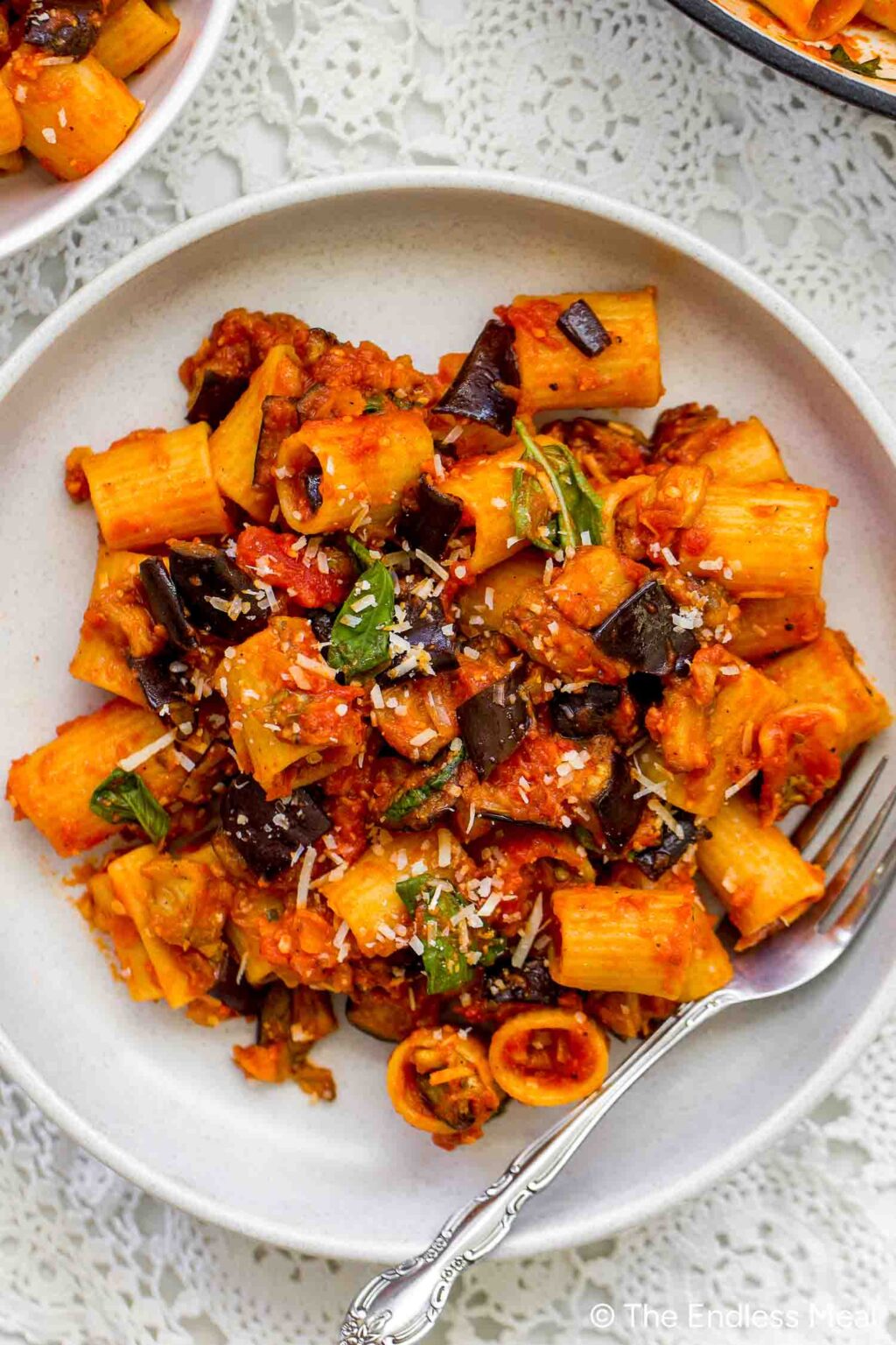A plate of Pasta alla Norma with rigatoni, rich tomato sauce, eggplant, basil, and grated cheese is served with a fork on a white lace tablecloth.