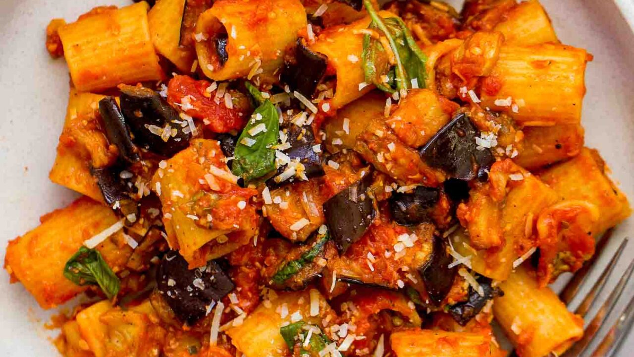 A plate of Pasta alla Norma with rigatoni, rich tomato sauce, eggplant, basil, and grated cheese is served with a fork on a white lace tablecloth.