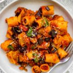 A plate of Pasta alla Norma with rigatoni, rich tomato sauce, eggplant, basil, and grated cheese is served with a fork on a white lace tablecloth.