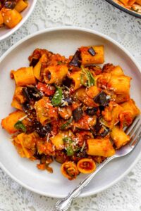 A plate of Pasta alla Norma with rigatoni, rich tomato sauce, eggplant, basil, and grated cheese is served with a fork on a white lace tablecloth.